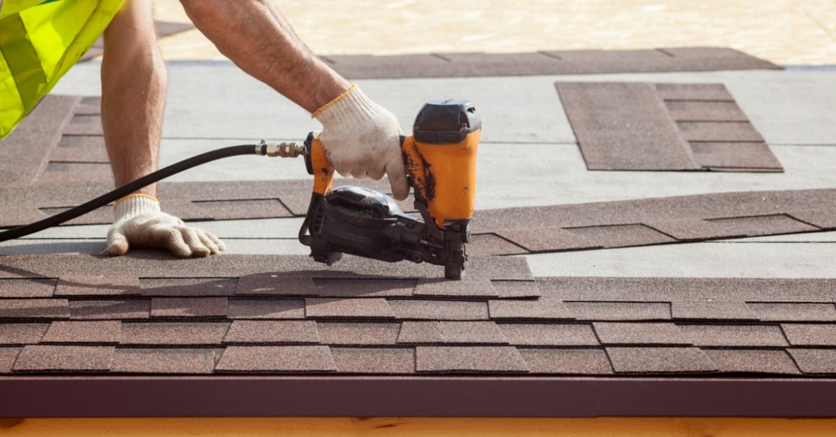 A worker in white gloves and a high-visibility vest fastening brown asphalt roof shingles with an orange nail gun.
